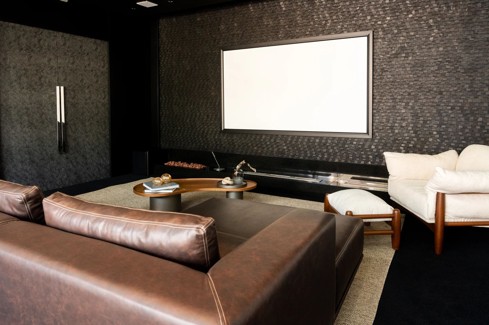 Ambiente de home theater com sofá em couro, poltrona clara e tela de projeção em parede texturizada